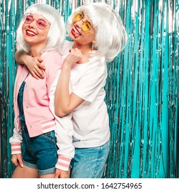 Two young sexy smiling hipster girls in white wigs and red lips.Beautiful trendy women in summer clothes.Carefree models posing on blue silver shiny tinsel background in studio.Show tongues in glasses