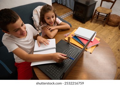 Two young siblings happily engaged with their laptop for learning and homework, surrounded by notebooks and study materials at home. - Powered by Shutterstock