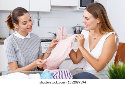 Two young smiling girlfriends looking new clothes at table and sharing 