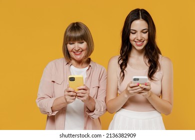 Two Young Smiling Happy Daughter Mother Together Couple Women In Casual Beige Clothes Hold In Hand Using Mobile Cell Phone Isolated On Plain Yellow Color Background Studio. Family Lifestyle Concept