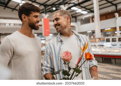 Two young smiling men are holding a bouquet of flowers while shopping together in a supermarket, enjoying their time together - Powered by Shutterstock