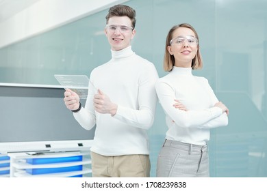 Two Young Smiling Programmers In Smart Casualwear And Goggles Standing On Background Of Large Interactive Display In Office