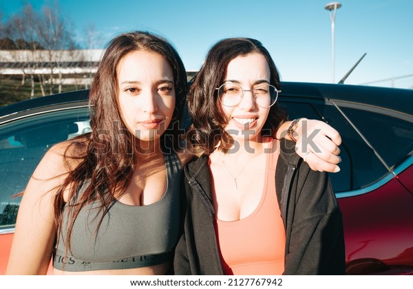 Two young sporty woman friends serious to camera\
before joining the gym, resting against car. Training losing weight\
with friends together smiling to camera happy. Friendship between\
sisters concept