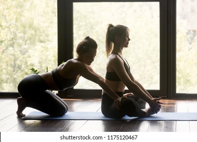 Two young sporty women practicing yoga, doing Butterfly exercise, baddha konasana pose, working out wearing sportswear, indoor full length, yoga studio, student training in sport club with instructor