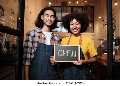 Two young startup barista partners with aprons stand at casual cafe door, letters on board and show open sign, happy and cheerful smiles with coffee shop service jobs, and new business entrepreneurs.