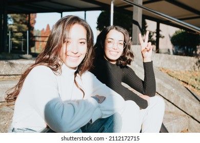 Two Young Students Outside The University Campus Sitting And Smiling To Camera While Doing The Victory Sign To Camera. Happy To Study, Bright Future And Hard Work At Class Concept. Modern Style