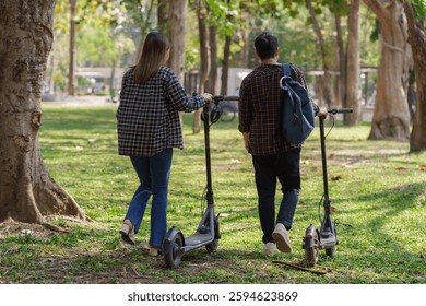 Two young students walking through a lush green park, pushing their electric scooters while enjoying a leisurely stroll under the warm sun, embracing a carefree day outdoors