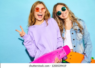 Two young stylish smiling blond women with penny skateboards. Models in summer hipster sport clothes posing near blue wall in studio. Positive girls going crazy