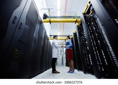 Two young technicians of data center consuting about working points at meeting while standing in aisle of storage room