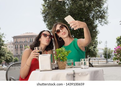 Two Young Tourist Girls Sit At A Bar Table While Taking A Selfie In Naples - Italy