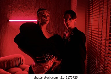 Two Young Vogue Dancers In Posh Stage Costumes Giving Performance In Studio Of Modern Night Club Lit With Dark Red Light Of Neon Lamps