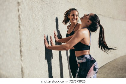 Two Young Woman Doing Stretching Workout Against A Wall And Smiling. Female Friends Exercising Together In Morning Outdoors.