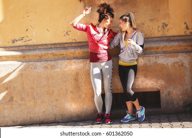 Two young woman standing against the wall on street and relaxing after jogging.Sharing the earphones and listening to music. - Powered by Shutterstock