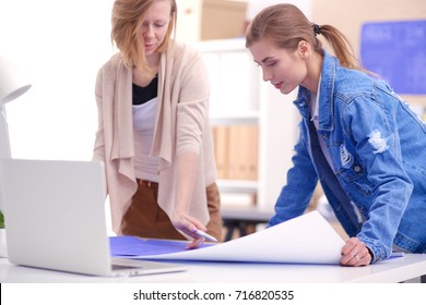 Two young woman standing near desk with instruments, plan and laptop.