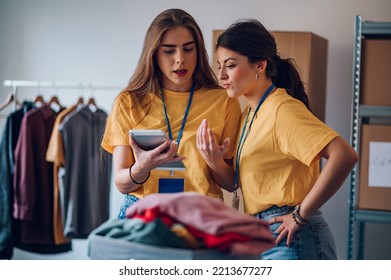 Two Young Woman Working In Charitable Foundation And Using A Digital Tablet. Happy Volunteer Separating Donation Stuff. Volunteers Sort Donations During Food Drive.