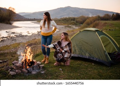Two young women bask in the campfire. Friends travel and relax comfortably on an autumn evening in the open air by the fire. Camping on green meadow on river and mountain background.