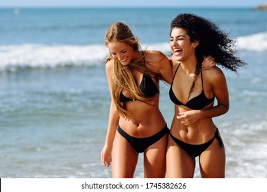 Zwei junge Frauen mit schönen Körpern in Badebekleidung an einem tropischen Strand. Hübsche, kaukasische und arabische Weibchen mit schwarzem Bikini, die am Ufer spazieren.