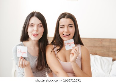 Two young women in bedroom at home sitting on bed holding paper with stigma word looking camera showing tongue smiling playful