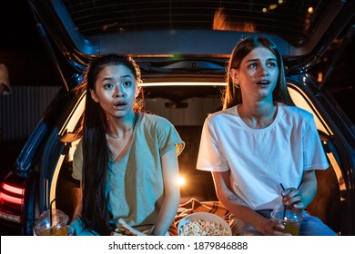 Two Young Women, Best Friends Looking Surprised While Sitting In Car Trunk, Having Snacks And Watching A Movie In A Drive In Cinema In The Evening. Entertainment, Friendship Concept. Horizontal Shot