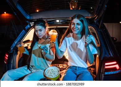 Two Young Women, Best Friends Looking Emotional While Sitting In Car Trunk, Having Snacks And Watching A Movie In A Drive In Cinema. Entertainment, Friendship Concept. Horizontal Shot