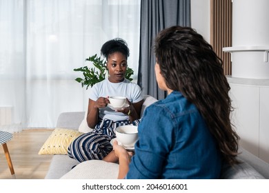 Two Young Women Best Friends Sitting On The Sofa At Home Drinking Tea Or Coffee Black African Is Complaining On Her Boyfriend Or Husband To White One How He Changed And Become Jealous, Selective Focus