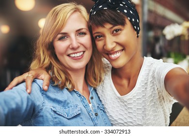 Two Young Women Best Friends Sitting Arm In Arm With Their Faces Close Together Smiling At The Camera, Multiethnic Couple