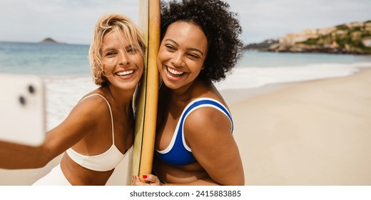 Two young women in bikinis, one holding a surfboard, take a beach selfie with a camera phone. They happily celebrate their friendship and create lasting memories on a fun surfing trip. - Powered by Shutterstock