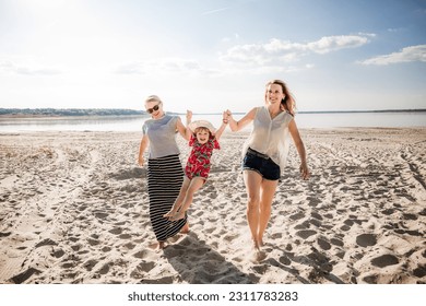 Two young women carry child along the beach. Family has fun. Mother and aunt support a little happy jumping girl. Summer vacation lifestyle. Smiling and laughing people. Good mood. Happy atmosphere. - Shutterstock ID 2311783283