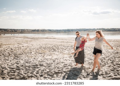 Two young women carry child along the beach. Family has fun. Mother and aunt support a little happy jumping girl. Summer vacation lifestyle. Smiling and laughing people. Good mood. Happy atmosphere. - Shutterstock ID 2311783285