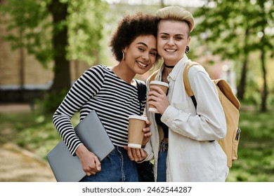 Two young women in casual attire holding coffee cups and a laptop while chatting in a park setting. - Powered by Shutterstock