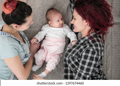 Two Young Women In Casual Clothes And With Pink Hair, A Lesbian Homosexual Couple, Lying On A Rug With A Child. Marriage, Adoption.