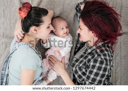 Two young women in casual clothes and with pink hair, a lesbian homosexual couple, lying on a rug with a child. Marriage, adoption.
