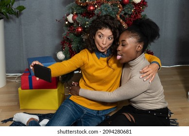 Two Young Women Celebrate Christmas And New Year's Eve Together By Joking And Taking Selfies With A Smartphone Goofy And Making Funny Facial Expressions In Front Of The Christmas Tree. Holidays Concep