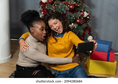 Two Young Women Celebrate Christmas And New Year's Eve Together By Joking And Taking Selfies With A Smartphone Goofy And Making Funny Facial Expressions In Front Of The Christmas Tree. Holidays Concep