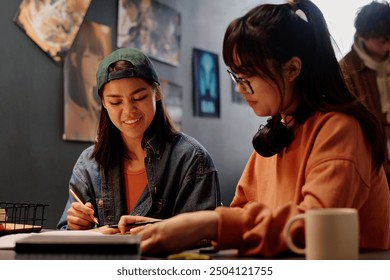 Two young women collaborating on creative project in casual setting with art posters on wall. One wears a cap while writing and other wears headphones while focusing on work - Powered by Shutterstock