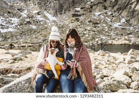 
Two young women, covered with a blanket, on a sunny winter morning enjoying a day on the mountain with their cute little dog taking themselves pictures with their smartphone. Lifestyle.