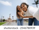 Two young women embracing and taking a selfie in plaza de armas, with the monument to liberty in the background, in trujillo, peru