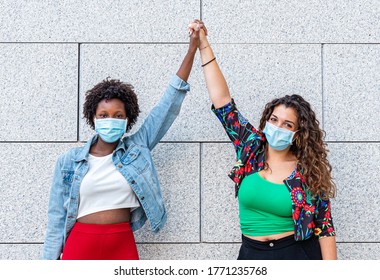 Two Young Women Friends, One White And One Black, Raise Their Fists Together As A Sign Of Sisterhood And Protest Against Racism, New Socialization With Protective Masks