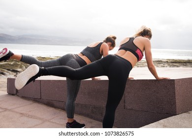 Two Young Women Getting Fit Together While Doing Leg Stretches In Sporty Clothing