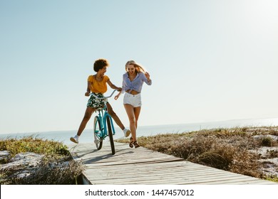 Two young women having fun with a bicycle at the beach. Woman running with friend riding a bike on boardwalk. - Powered by Shutterstock