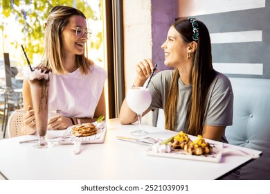 Two young women are having a snack in a modern cafe. The friends are having sweets, milkshakes and ice cream cups for dessert. Concept of friends meeting in a cafe and having sweets. - Powered by Shutterstock