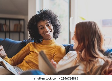 two young women at home, a couple of lesbian sitting in sofà in living room, reading books and having moments of relax, smiling african woman looking her girlfriend - Powered by Shutterstock