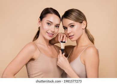 Two Young Women With Natural Visage Posing With Tube Of Concealer Isolated On Beige