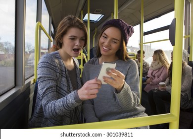 Two Young Women On A Bus. They Are Both Looking At Something On A Smart Phone. There Are People On The Bus In The Background.