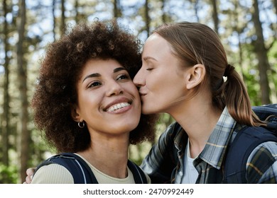 Two young women, one with curly hair and one with a ponytail, are hiking in the woods. They are both smiling and happy. The woman with the ponytail is kissing the other woman on the cheek. - Powered by Shutterstock