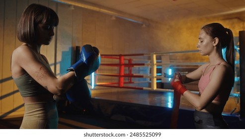 Two young women preparing for a boxing match by wrapping bandages on their hands in front of a boxing ring, emphasizing fitness, training, and active lifestyle in a modern gym with colored lighting - Powered by Shutterstock