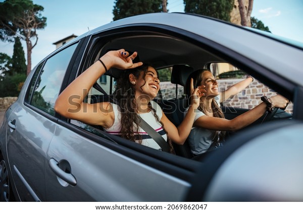 Two young women sing a song on the radio and dance
in the car on a day trip in the summer - Best friends having fun
together driving around the countryside - Smiling millennial in a
relaxing moment