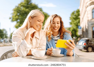 Two young women, sisters, best friends showing news social media photos on smart phone cellphone sitting in cafe and drinking coffee during breakfast. Fashion, beauty, blogging, tourism. - Powered by Shutterstock