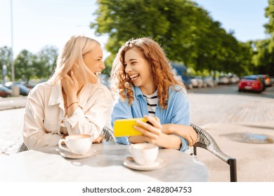 Two young women, sisters, best friends showing news social media photos on smart phone cellphone sitting in cafe and drinking coffee during breakfast. Fashion, beauty, blogging, tourism. - Powered by Shutterstock