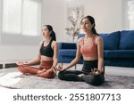 Two young women are sitting on a rug in a living room, practicing yoga and meditating in the lotus position, promoting a healthy lifestyle and mindfulness